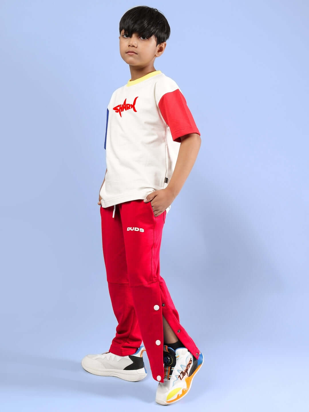 Boy wearing a shark fish printed oversized off-white t-shirt, paired with red pants, against a blue background, showcasing stylish attire.