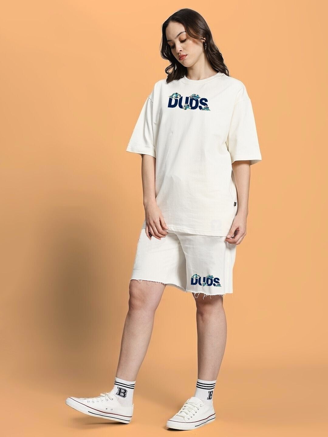 Woman wearing WOMEN CLOUD PRINTED RELAXED FIT SHORTS OFF WHITE, paired with casual T-shirt and sneakers, standing against a peach background.