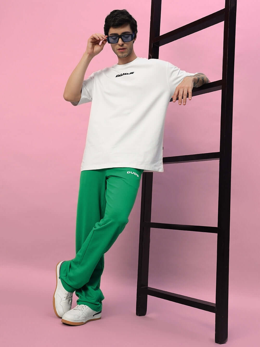 Model wearing NEW ERA RELAXED FIT JOGGERS in green, styled with a white t-shirt and sunglasses, leaning on a ladder.