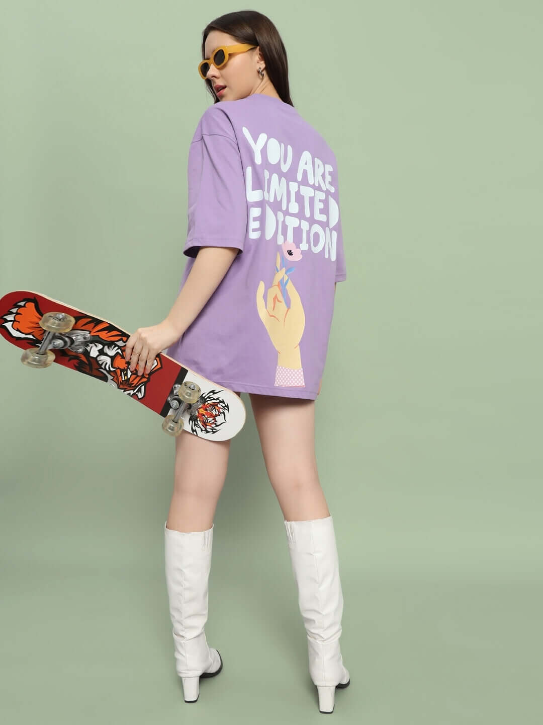 Woman in lavender oversized t-shirt with skater graphic, holding a skateboard, wearing white boots and sunglasses, back view.