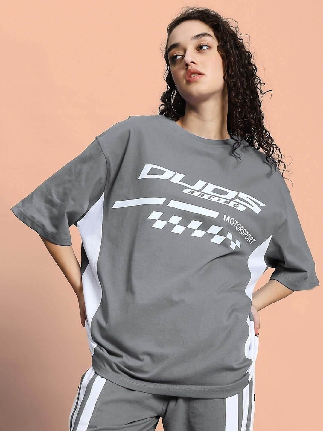 Woman wearing oversized grey t-shirt with printed graphics, hands on hips, styled on a peach background.