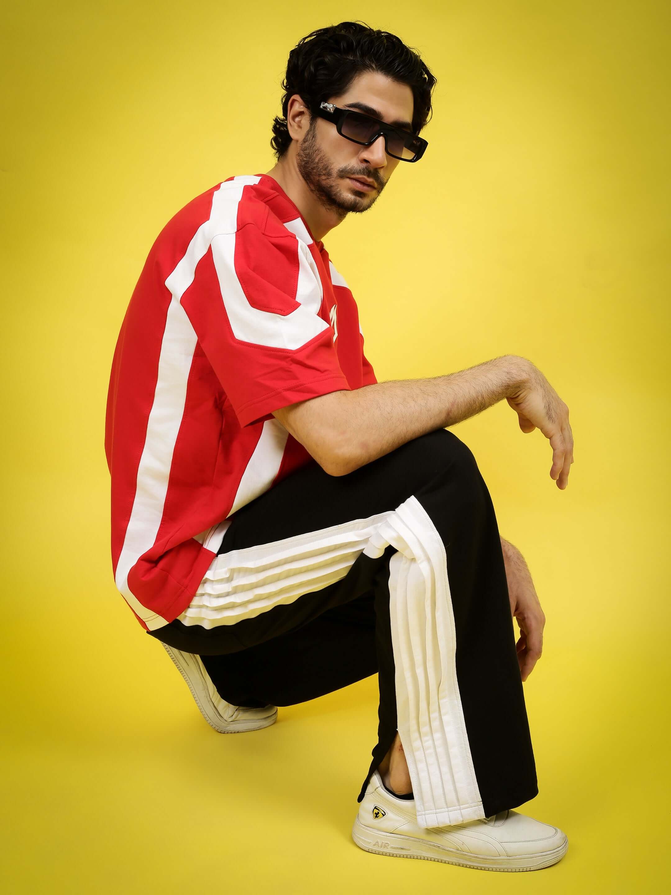 Male model in red and white shirt with black and white joggers, wearing sunglasses, posing on a yellow background