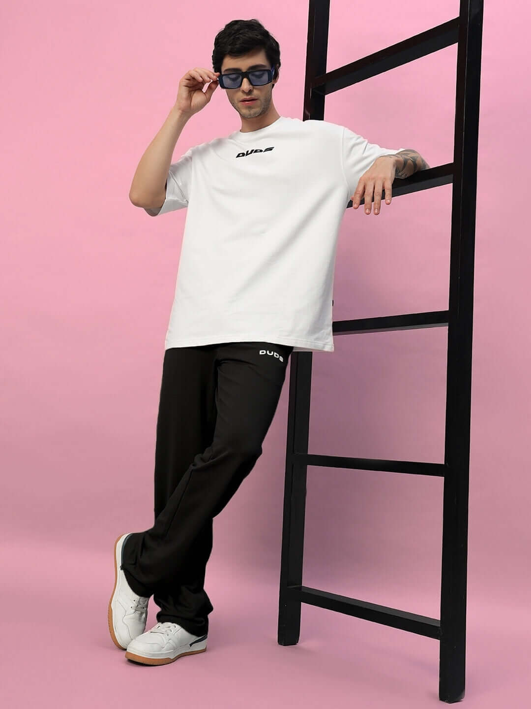Man wearing New Era Relaxed Fit Joggers in black, paired with a white shirt and sunglasses, leaning confidently against a ladder.