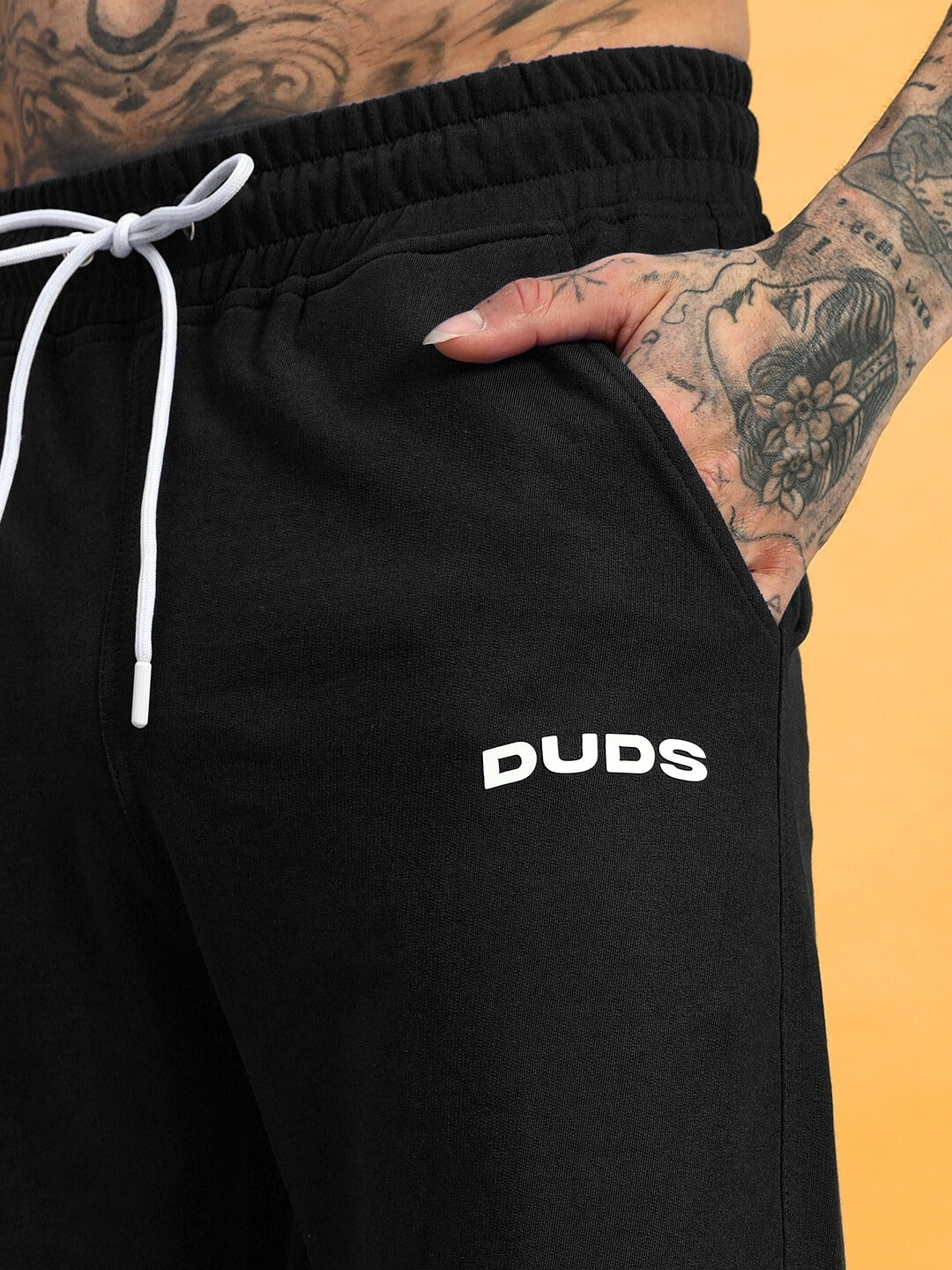 Close-up of black Hershey side slit joggers by DUDS, featuring white drawstrings and logo detail, modeled by a tattooed individual.