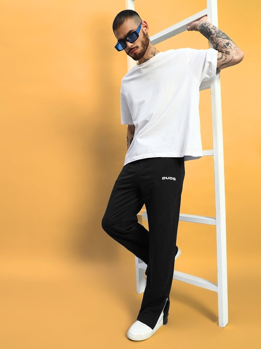 Man wearing Hershey side slit joggers in black by DUDS; casual style with white t-shirt and sunglasses, leaning on a ladder.