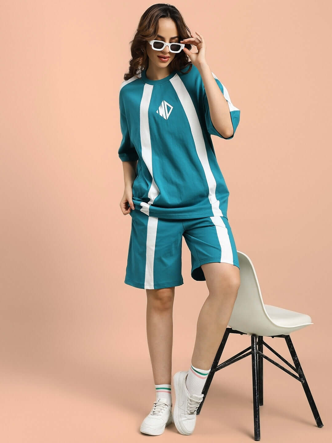 Model wearing teal WOMEN'S WIDELINE REGULAR FIT SHORTS with matching top, posing against a peach background in white sneakers and sunglasses.