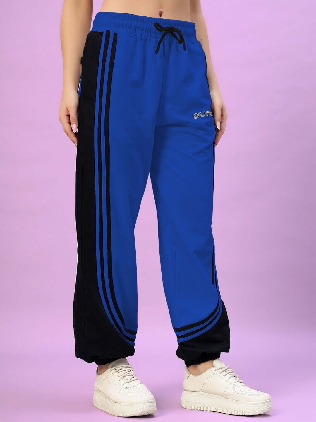 Woman wearing blue and black contrast joggers, featuring a drawstring waist, styled with white sneakers, on a pink background.
