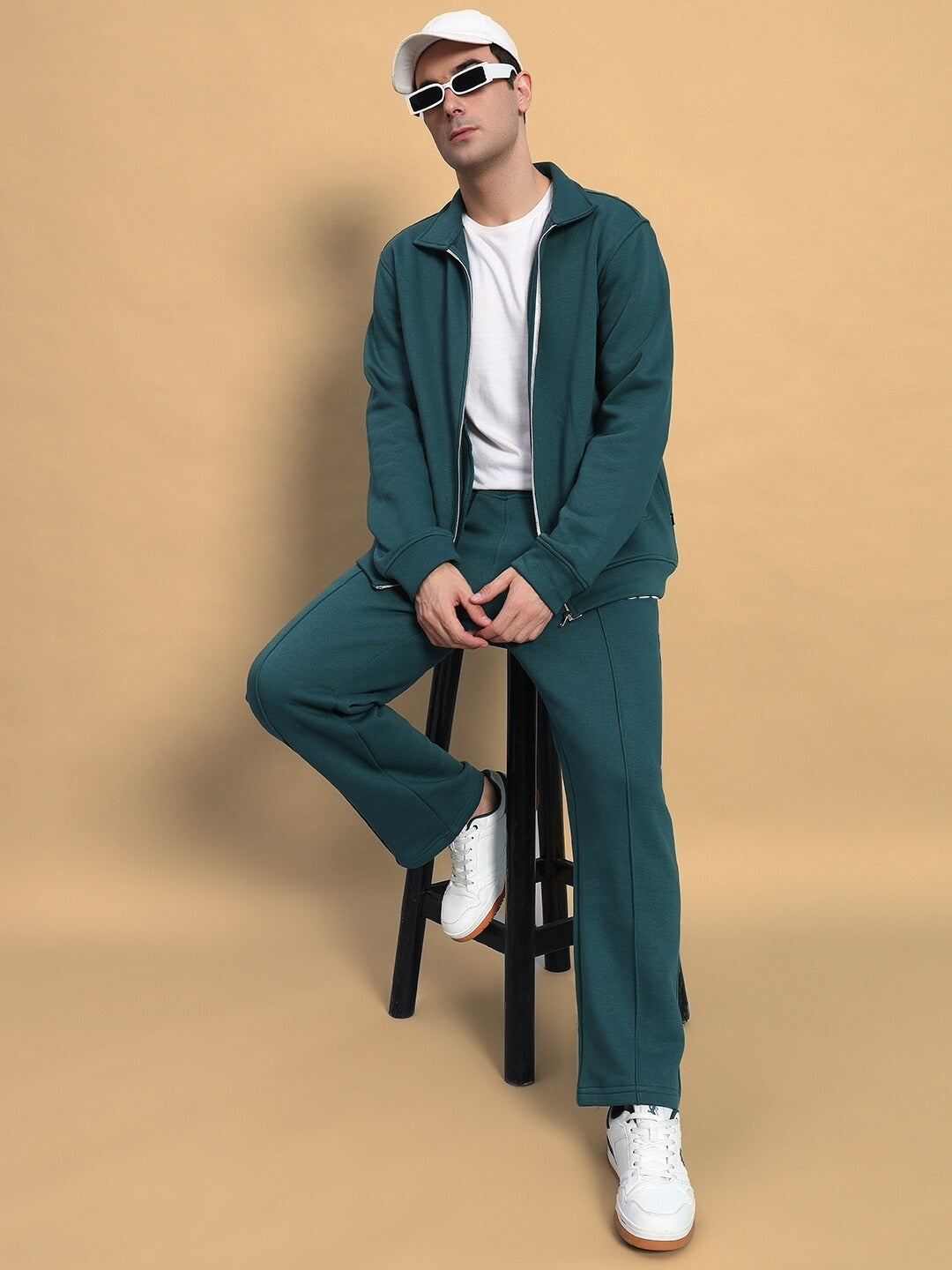 Man wearing teal green shaggy fleece jogger outfit, sitting on stool with white sneakers and hat, against beige background.