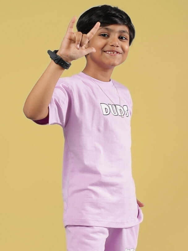 Smiling child wearing a lilac 'DUDS' wolf pack T-shirt for boys and girls, showing a playful hand gesture against a yellow background.