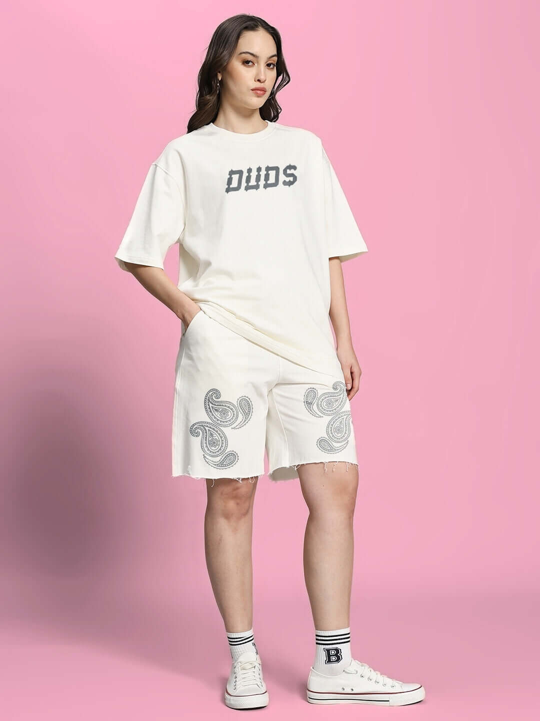 Woman wearing off-white Messi paisley print shorts and DUDS T-shirt, standing on pink background, showcasing casual fashion.
