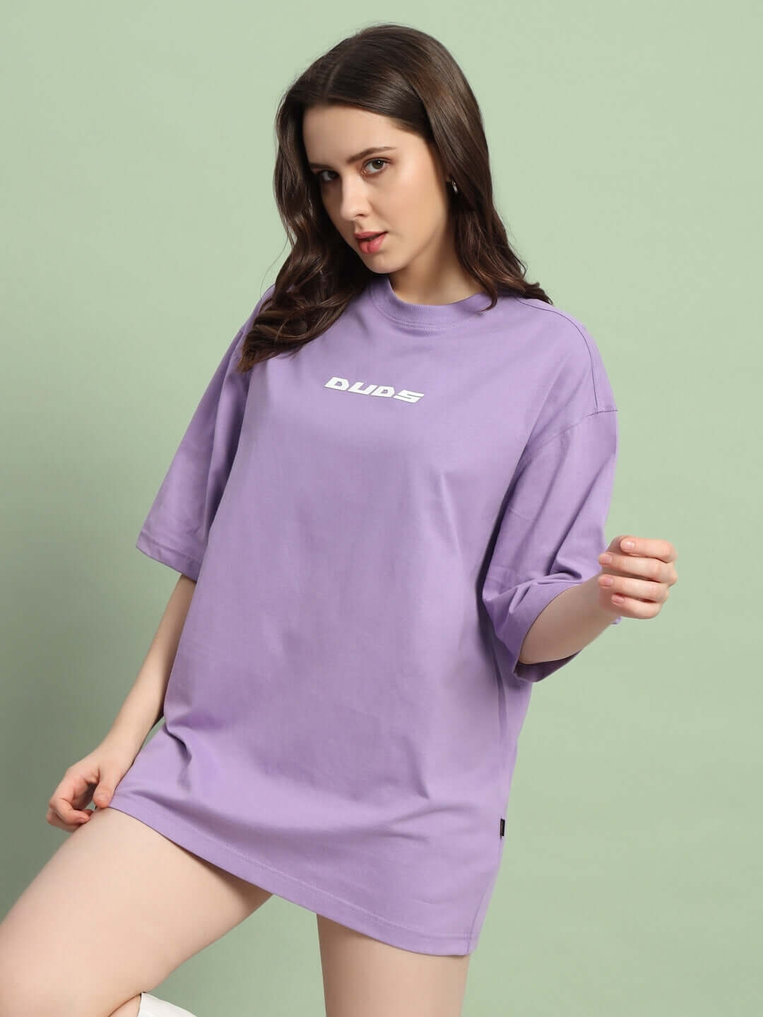 Woman wearing lavender oversized limited edition t-shirt by DUDS, showcasing relaxed fit and casual style against a green background.