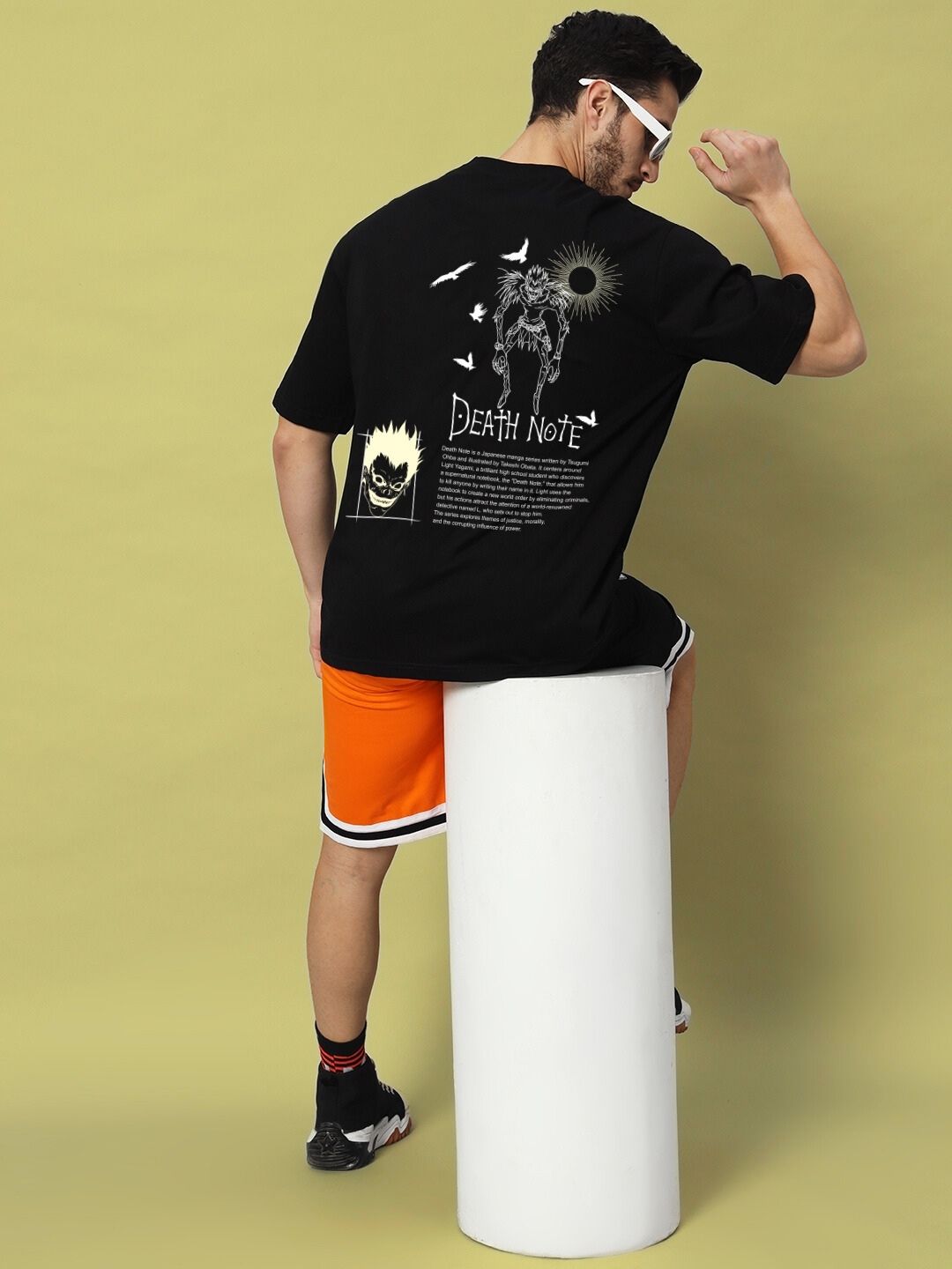 Rear view of man wearing FOLLOW LIFE OVERSIZED T-SHIRT BLACK featuring 'Death Note' design, sitting on a white cylinder against olive backdrop.