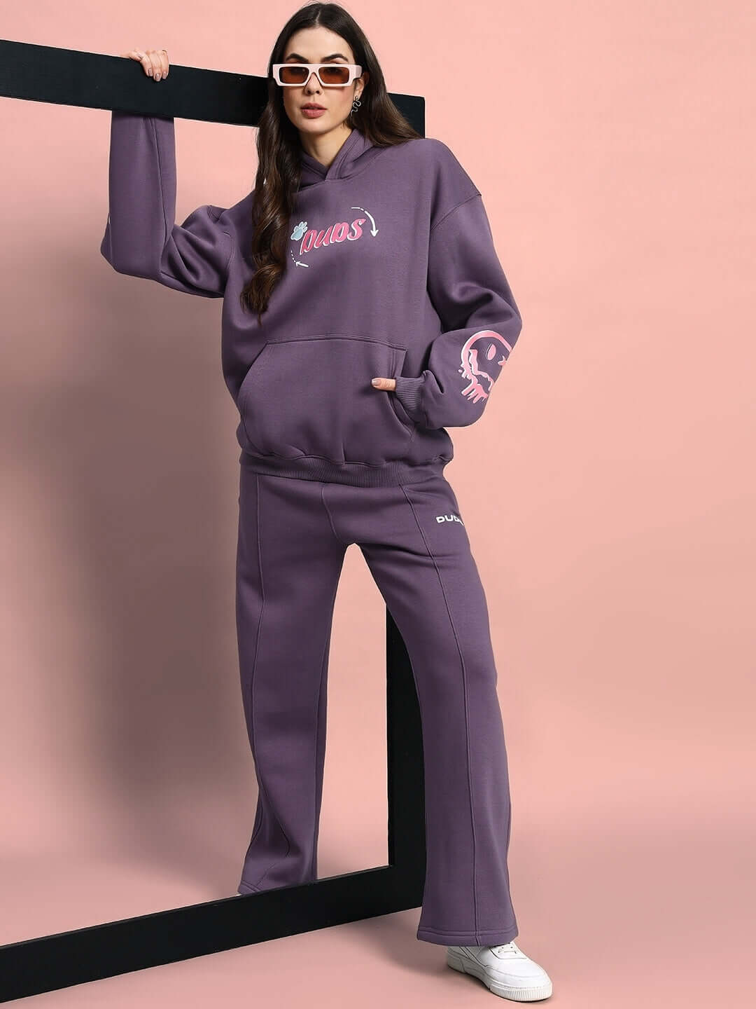 Woman in violet fleece jogger and hoodie set, wearing sunglasses, standing against a pink background.