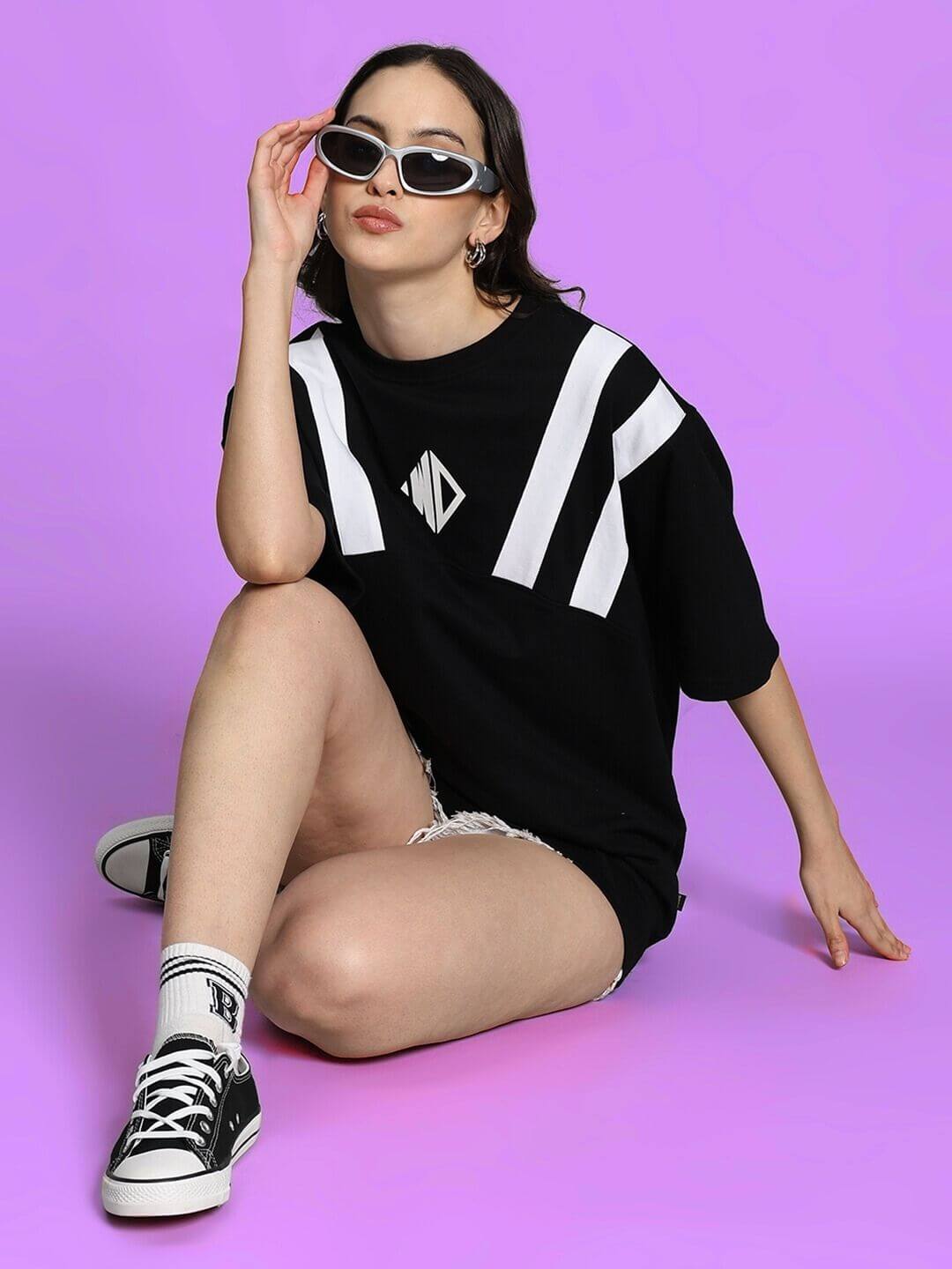 Model wearing Women's Alonzo oversized black T-shirt, styled with black sneakers and white socks, posing against purple background.