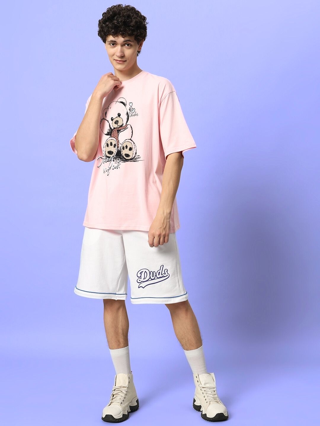 Young man wearing pink CUTE BEAR OVERSIZED T-SHIRT with white shorts and sneakers, standing against a light purple background.