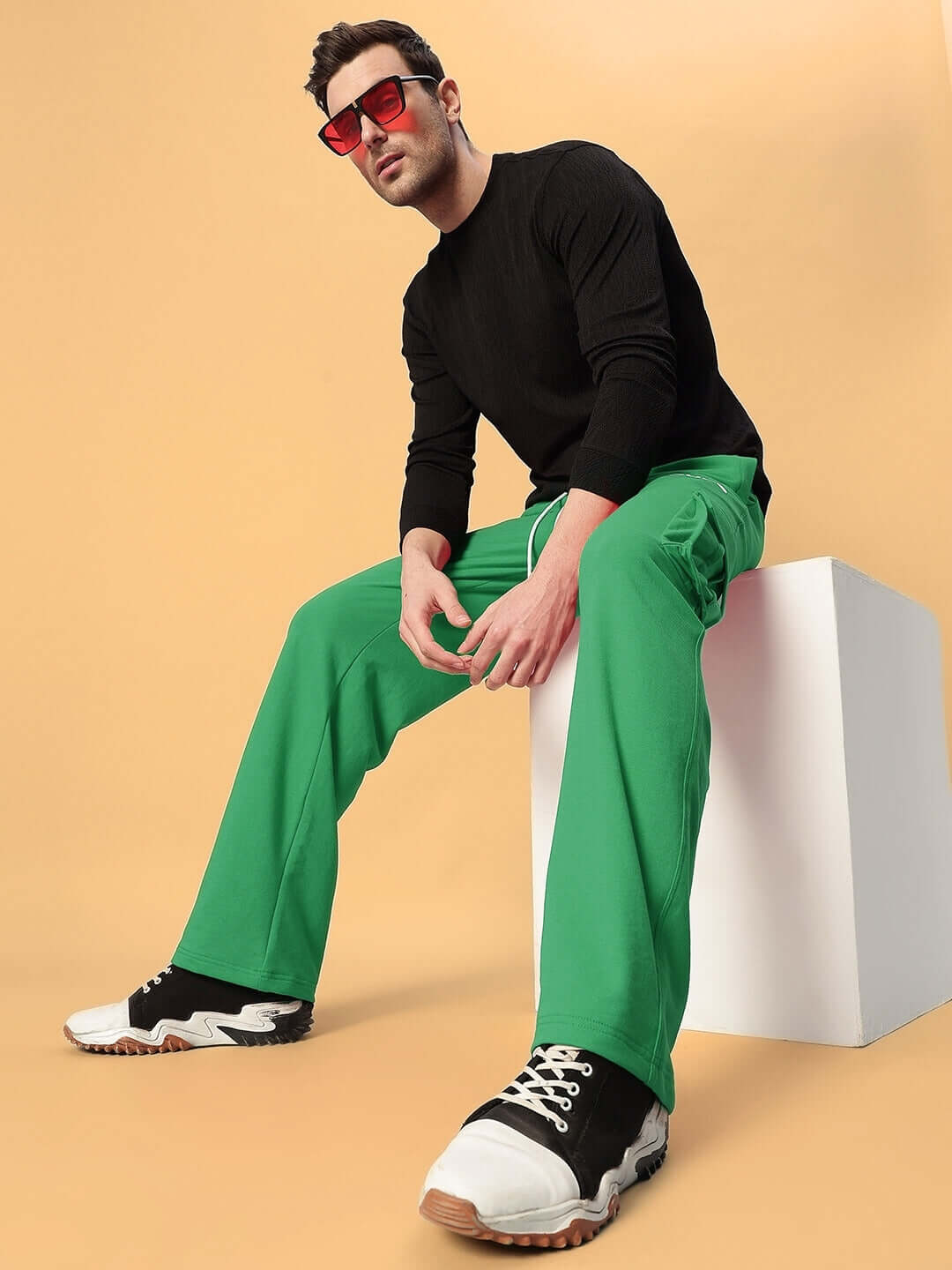Model wearing JESS 5 Pocket Relaxed Fit Joggers in green, paired with black top and sneakers, seated on a white cube against beige backdrop.