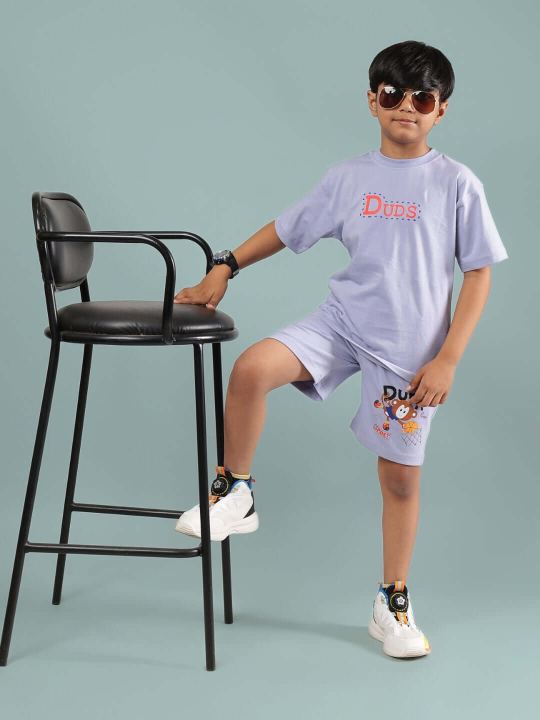 Child wearing lavender Sammy T-shirt by DUDS, standing by a chair, showcasing stylish unisex fashion for boys and girls.