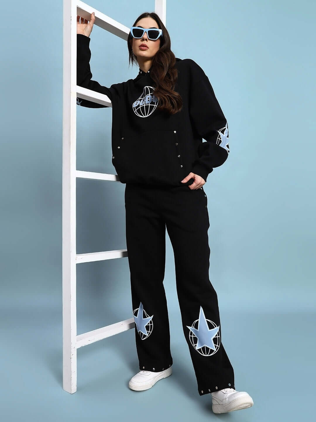 Woman wearing black embroidered fleece jogger outfit with sunglasses, leaning on a white ladder, blue background
