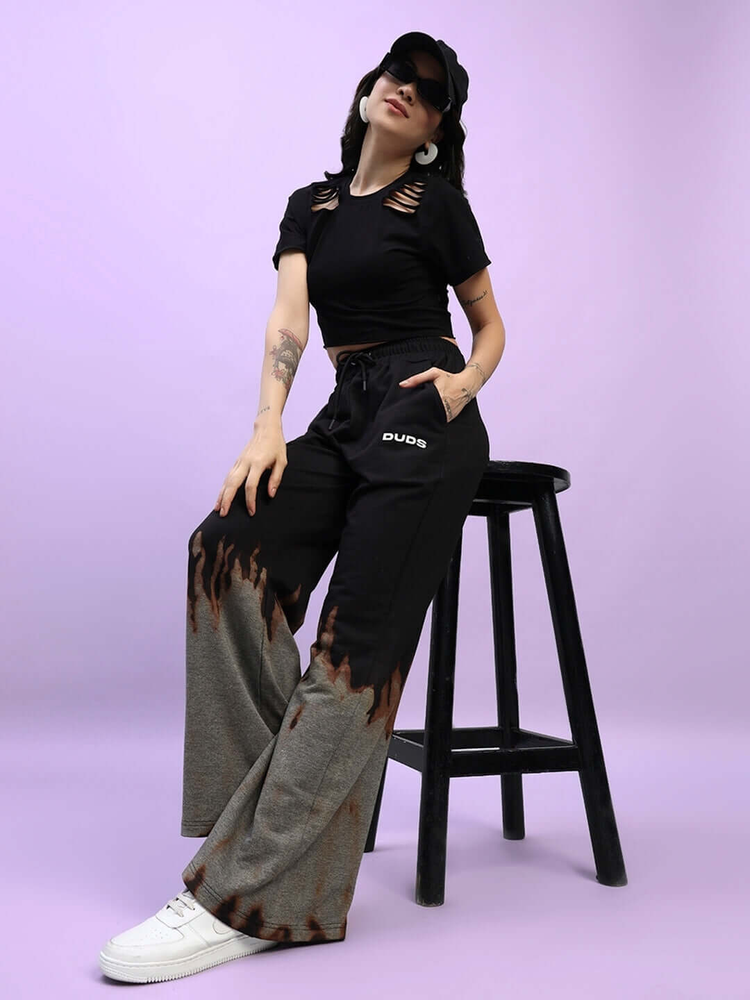 Model wearing women's fire flame baggy tie-dye jogger in black by DUDS, sitting on a stool with a casual style.