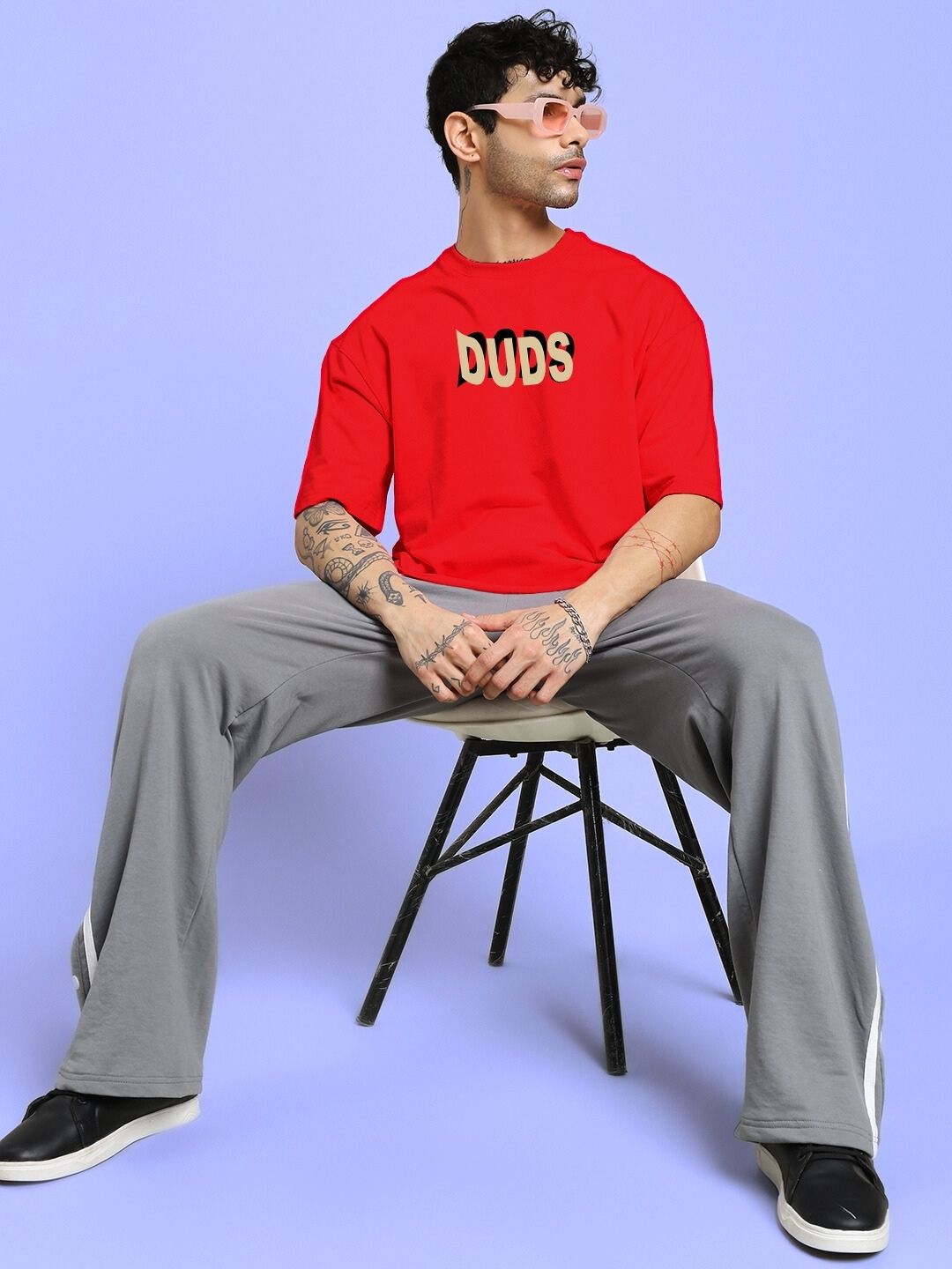 Model wearing KING OF SNAKE FUTURE PRINTED OVERSIZED T-SHIRT RED sitting stylishly on chair with gray pants and sunglasses.