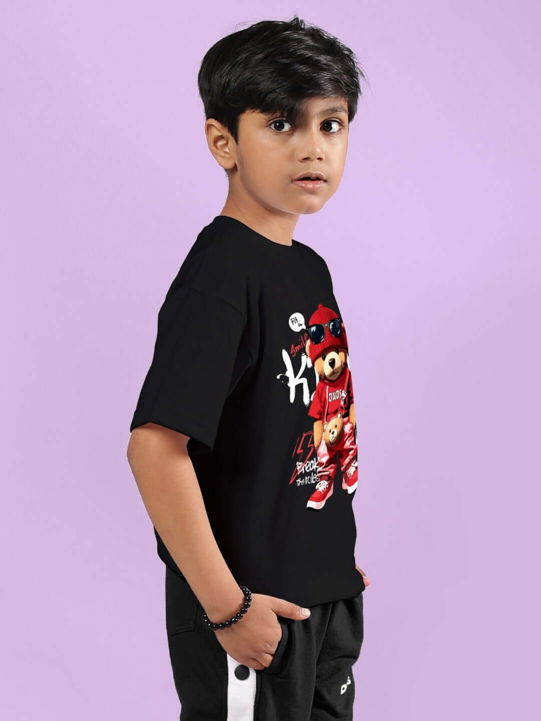 Young boy wearing a black hip hop teddy t-shirt, perfect for boys and girls, made by DUDS, standing against a pastel background.