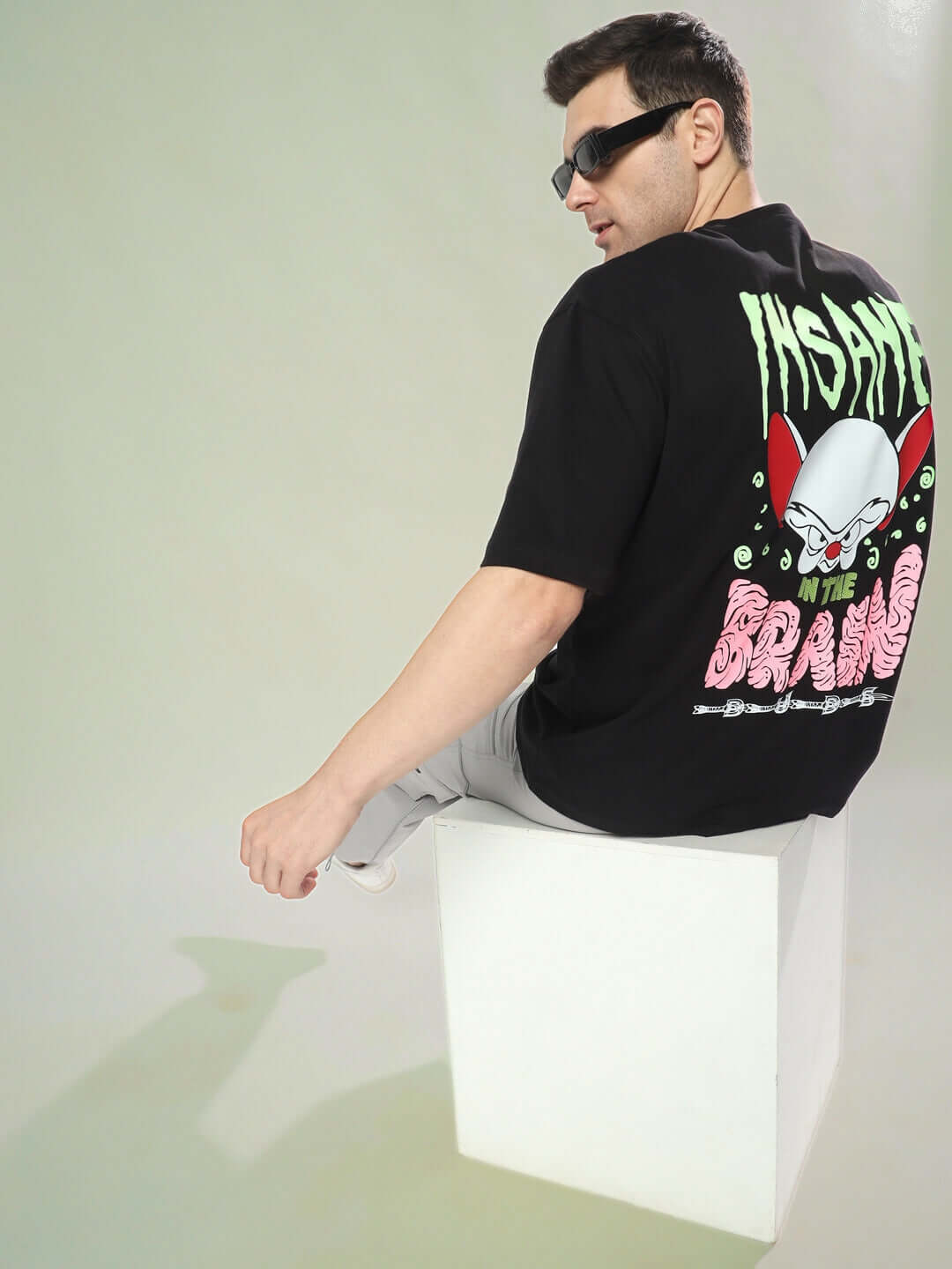 Man wearing INSANE BRAIN OVER-SIZED T-SHIRT BLACK sitting on a white block, showcasing back design graphics.