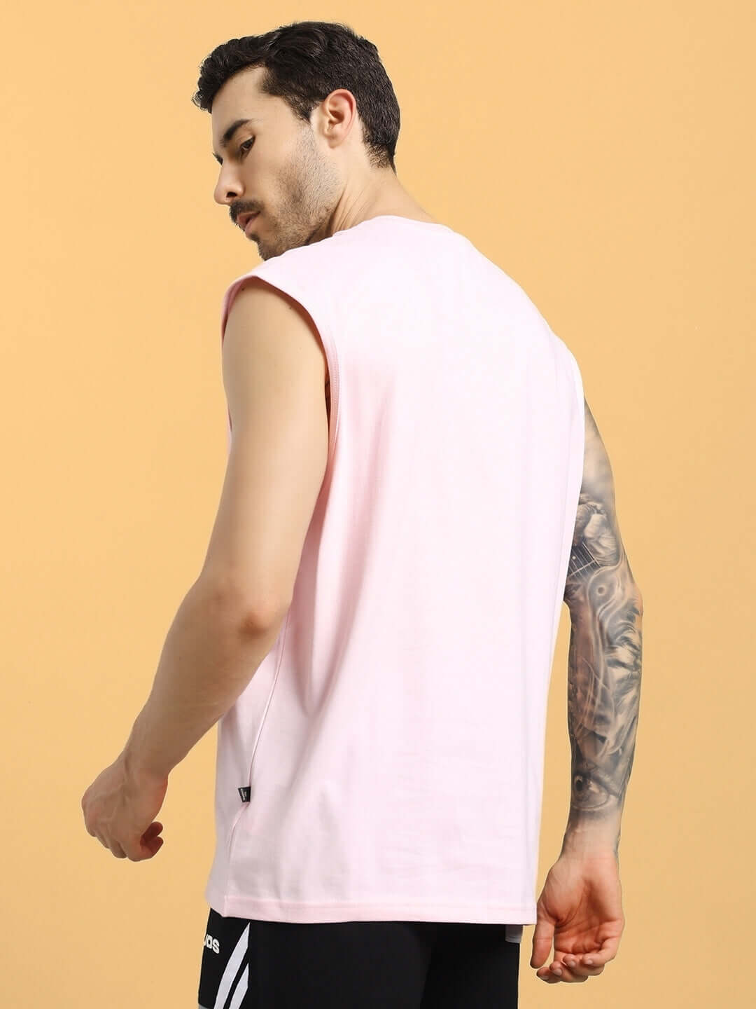 Back view of a man wearing a pink, sleeveless t-shirt with Panther graphic, featuring tattoos on his arm against a peach background.