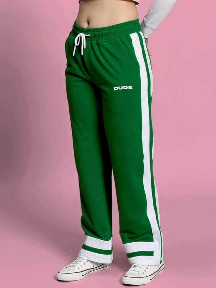 Woman wearing green joggers with side stripes, DUDS logo, standing against pink background.