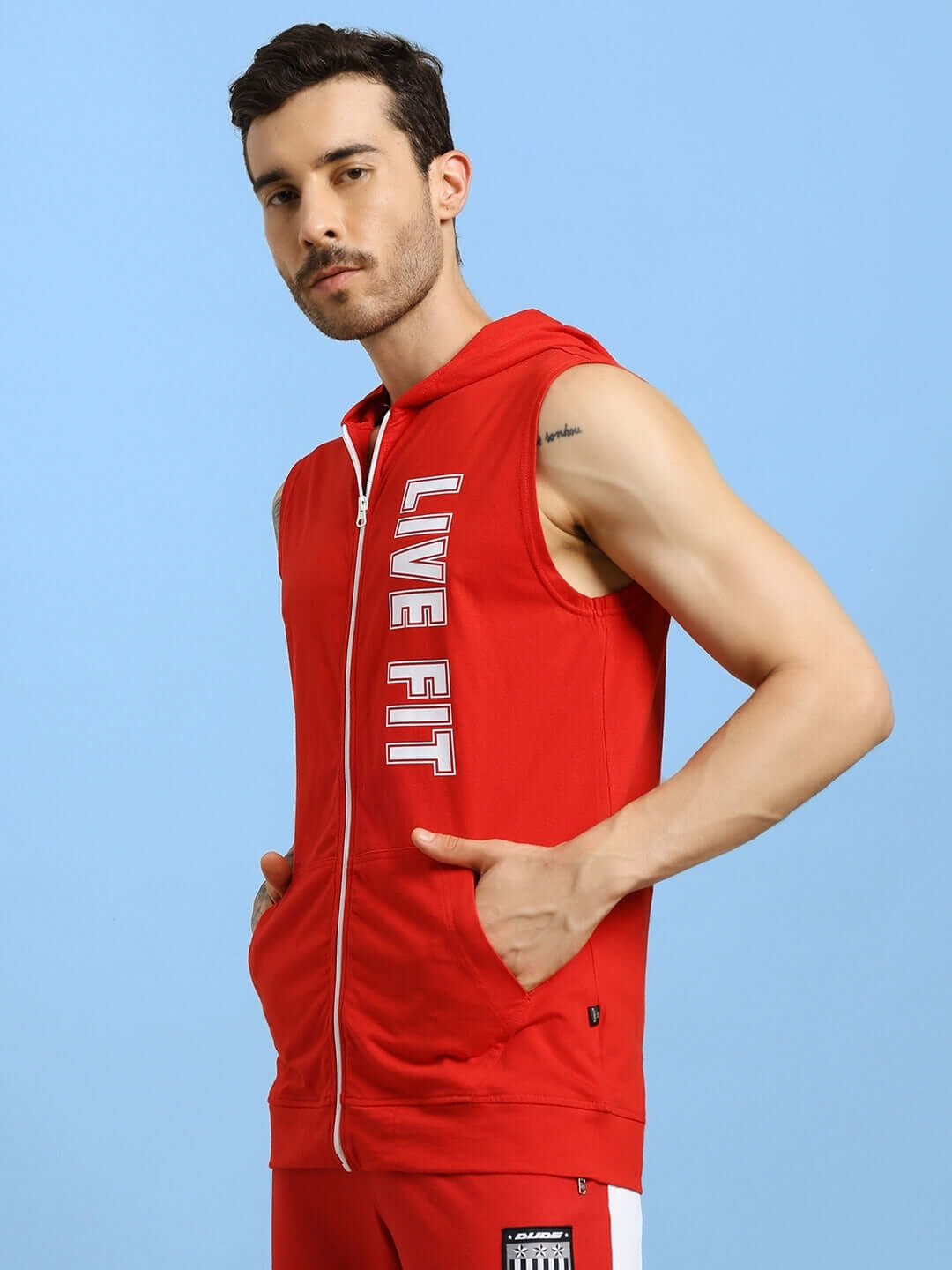 Man wearing DECENT LIVE FIT red hooded gym t-shirt with "Live Fit" text, showcasing sleeveless design and pockets, against a blue background.