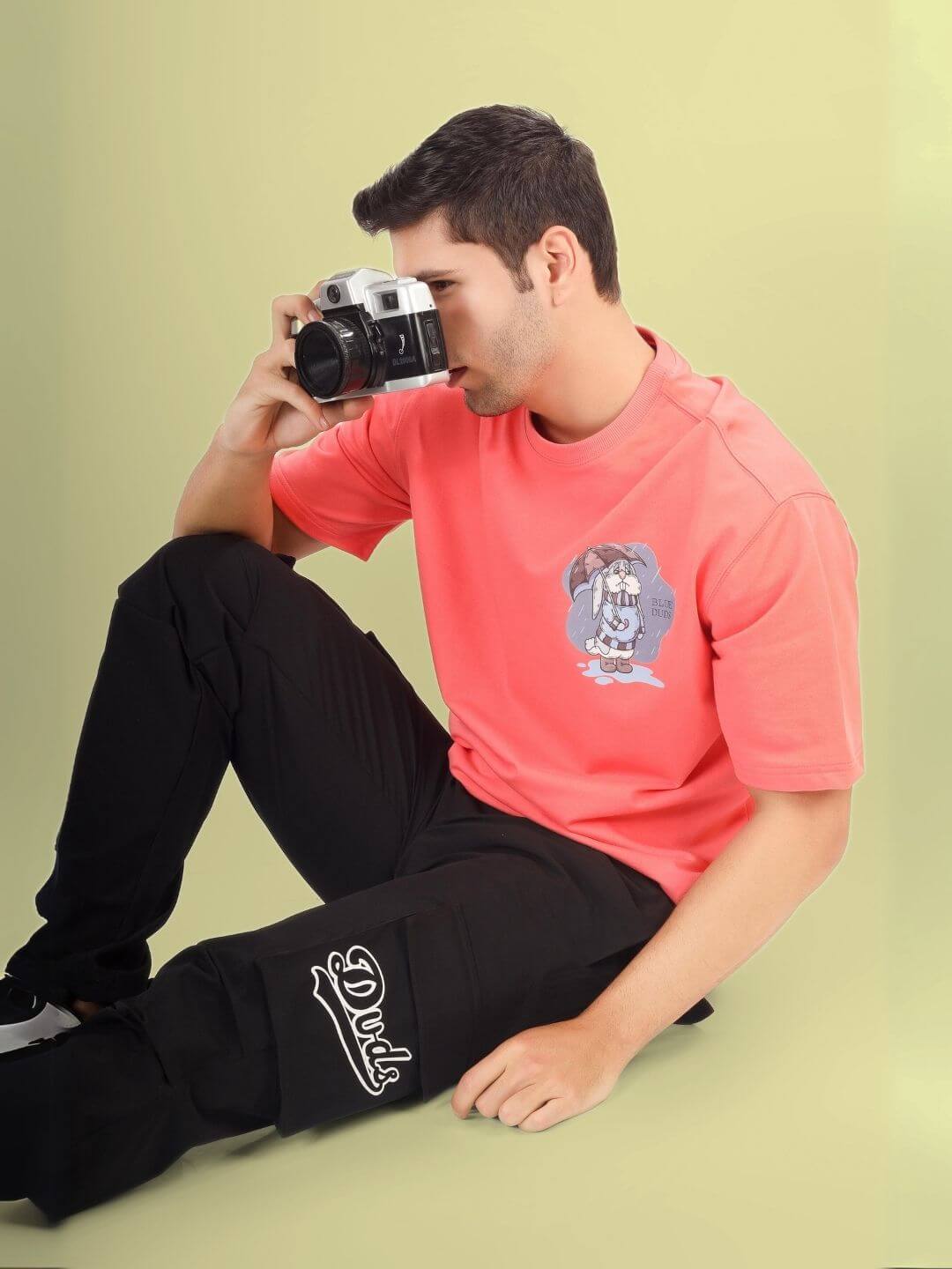 Man wearing BLUE LAPIN OVER-SIZED T-SHIRT PANTONE PEACH and black pants, holding a camera.