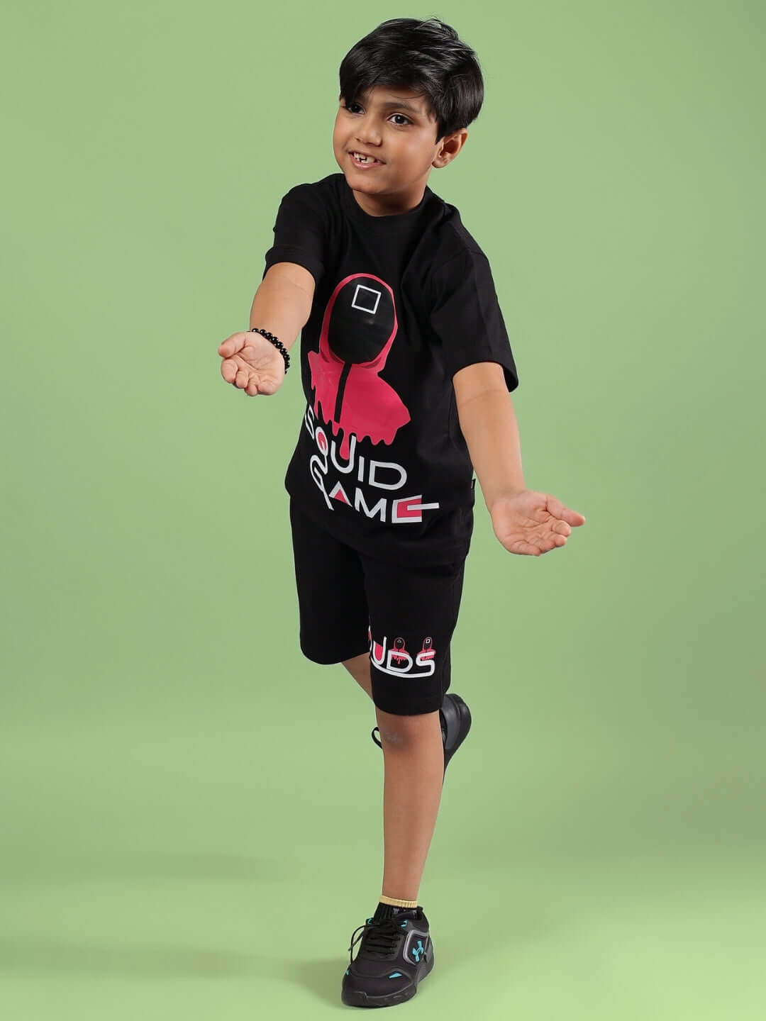 Child wearing black Squid Games co-ord set, featuring t-shirt and shorts with pink graphic, standing on green background.