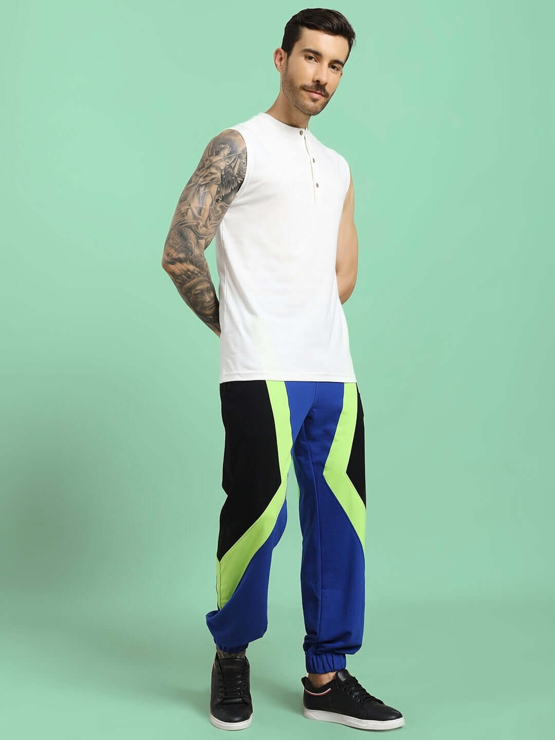 Man wearing a sleeveless white shirt paired with blue and green joggers, standing against a mint green background.