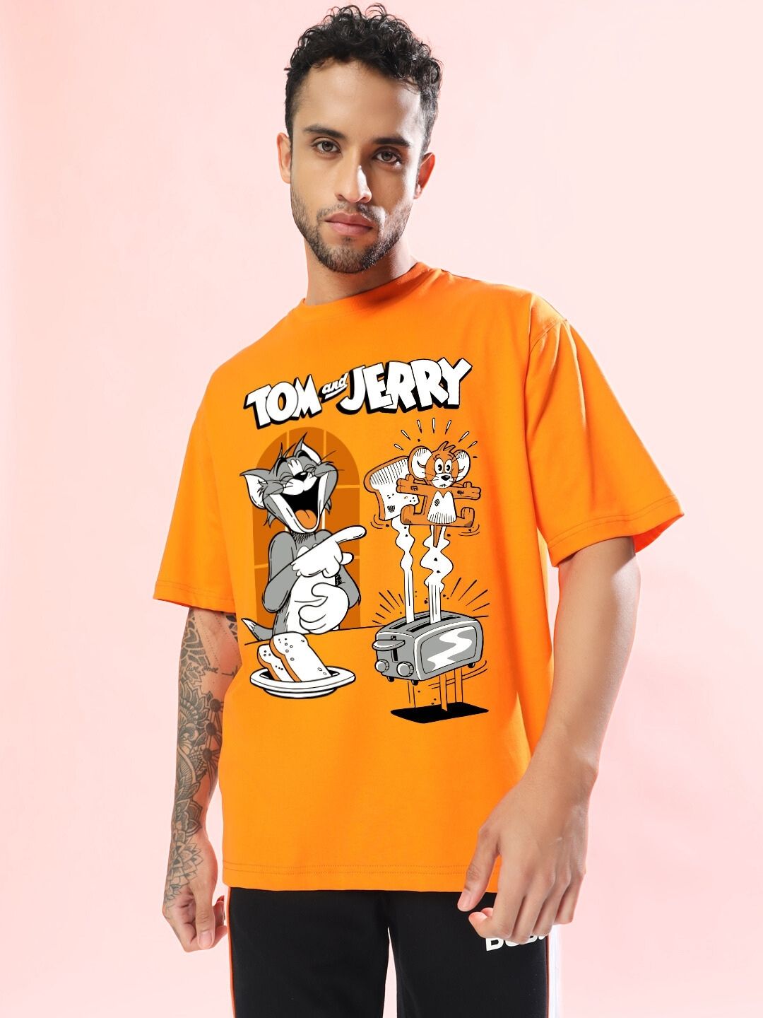 Man wearing TOM AND JERRY TOAST OVERSIZED T-SHIRT ORANGE with cartoon graphic print, standing against a pink backdrop.
