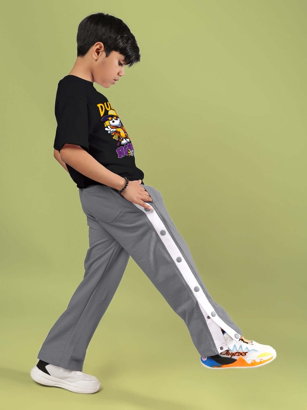 Young boy wearing black Snoopy Rimix t-shirt by DUDS, paired with gray pants and colorful sneakers, standing against a green background.