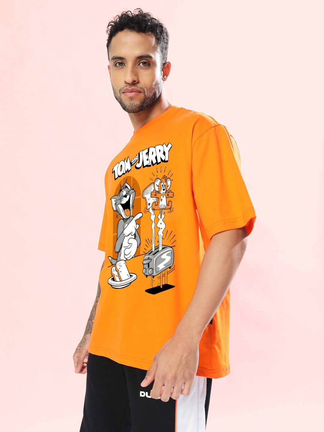 Model wearing TOM AND JERRY TOAST OVERSIZED T-SHIRT ORANGE, featuring cartoon characters, on a light pink background.