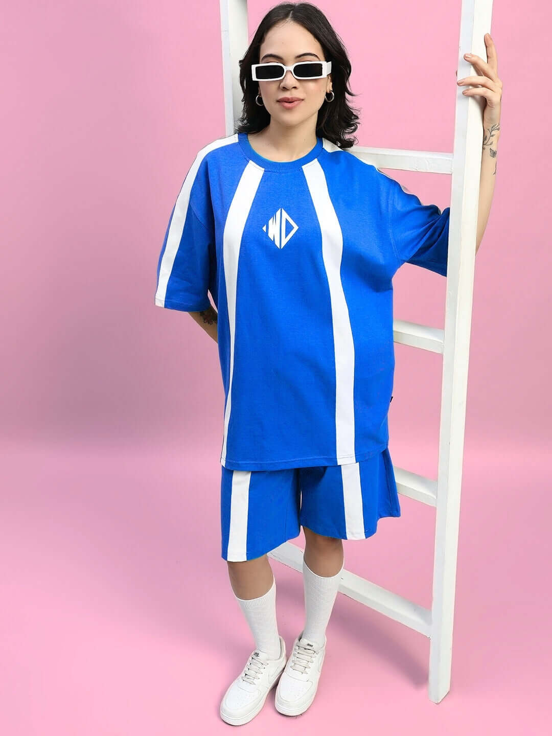 Model wearing blue women's wideline shorts and matching top, standing against a pink background with a white ladder.
