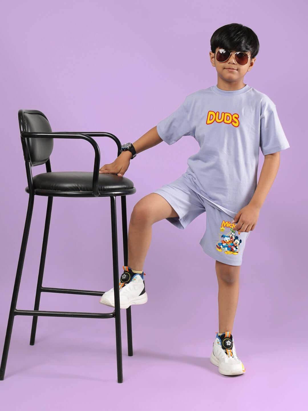 Child wearing lavender Mickey Dingo T-shirt and shorts with sunglasses, posing next to a black chair on a purple background.