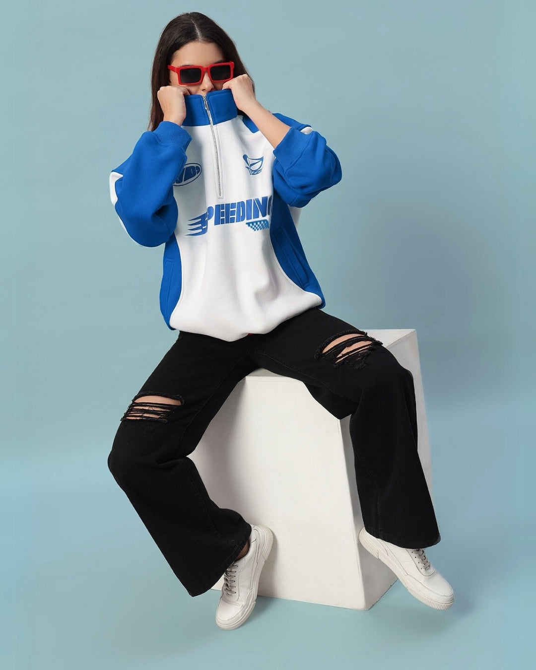 Woman wearing Women's Speeding Colorblock Jacket in royal blue with sporty design, ripped black jeans, and seated on a white block.