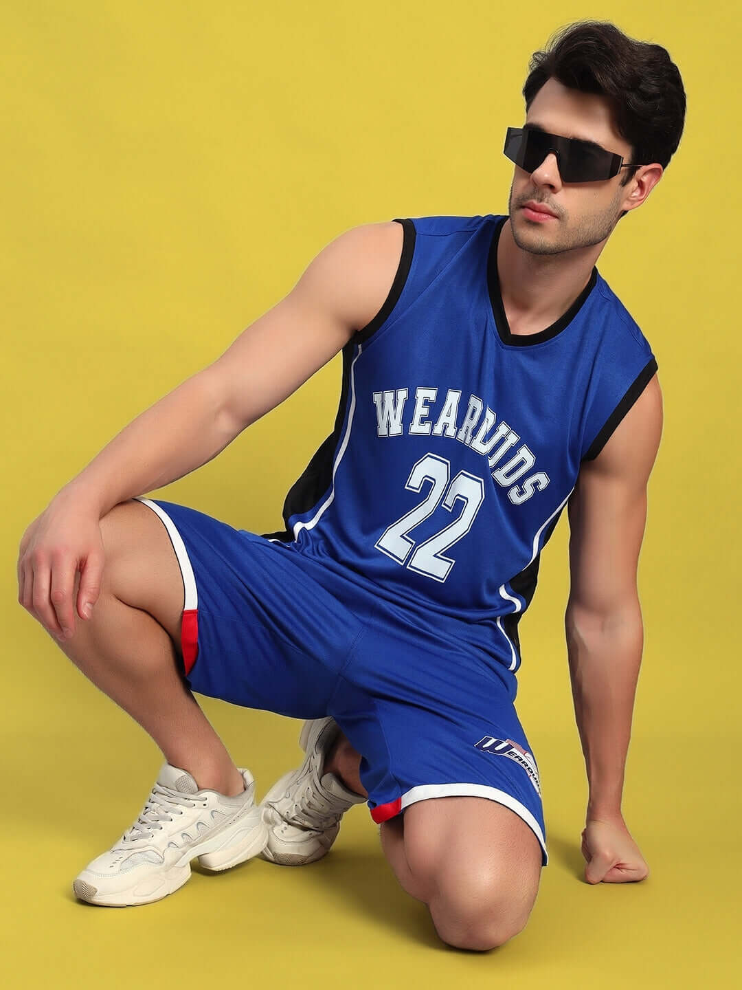 Man wearing a blue basketball jersey and shorts set with sunglasses, kneeling on a yellow background.