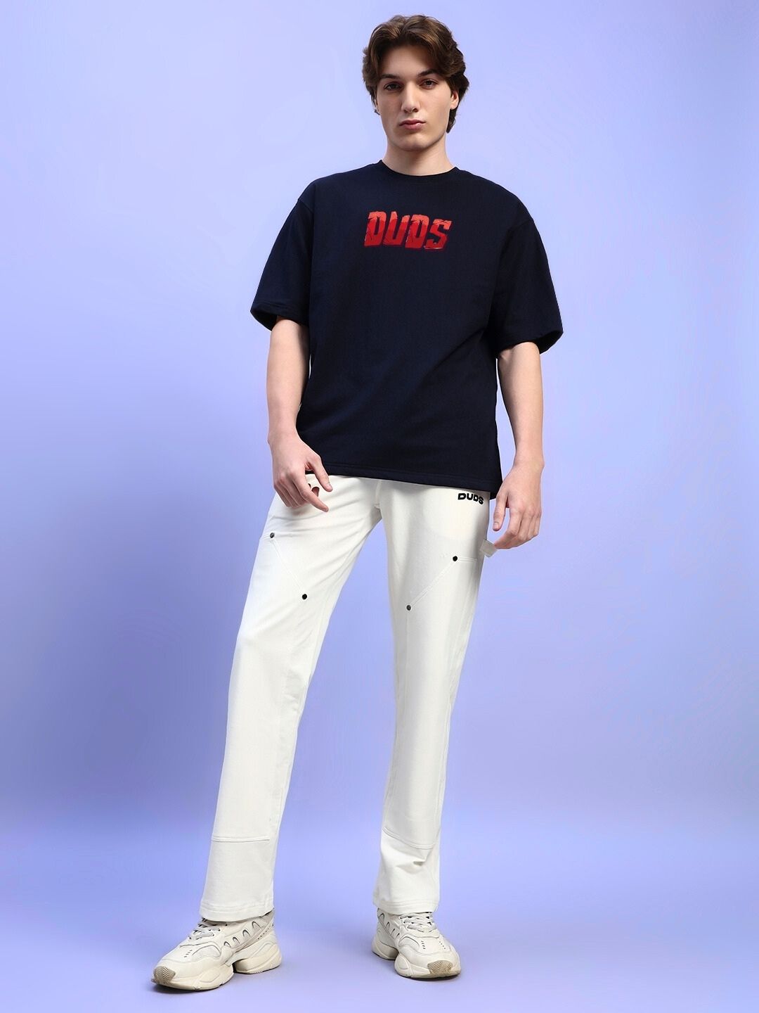 Model wearing AMPERES PRINT OVERSIZED T-SHIRT BLACK paired with white trousers and sneakers, standing against a light purple background.