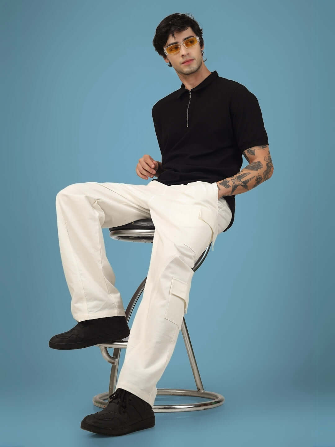 Model wearing 6 Pocket Cotton Twill Cargo Pant in white, sitting on stool against blue background, showcasing style and utility.