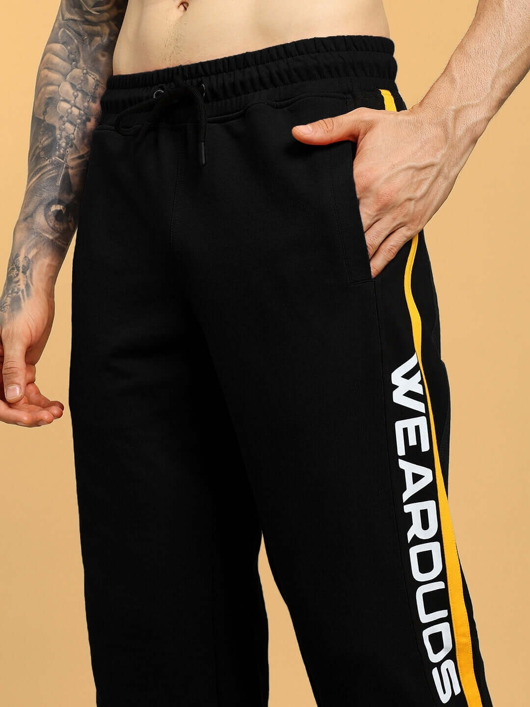 Close-up of black relaxed-fit joggers by DUDS with yellow stripe and 'WEARDUDS' text, featuring elastic waistband and stitching details.