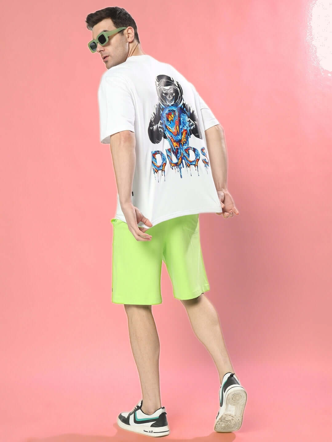 Man wearing MELTING EARTH OVER-SIZED T-SHIRT WHITE with bold graphic on back, paired with neon green shorts, against pink background.