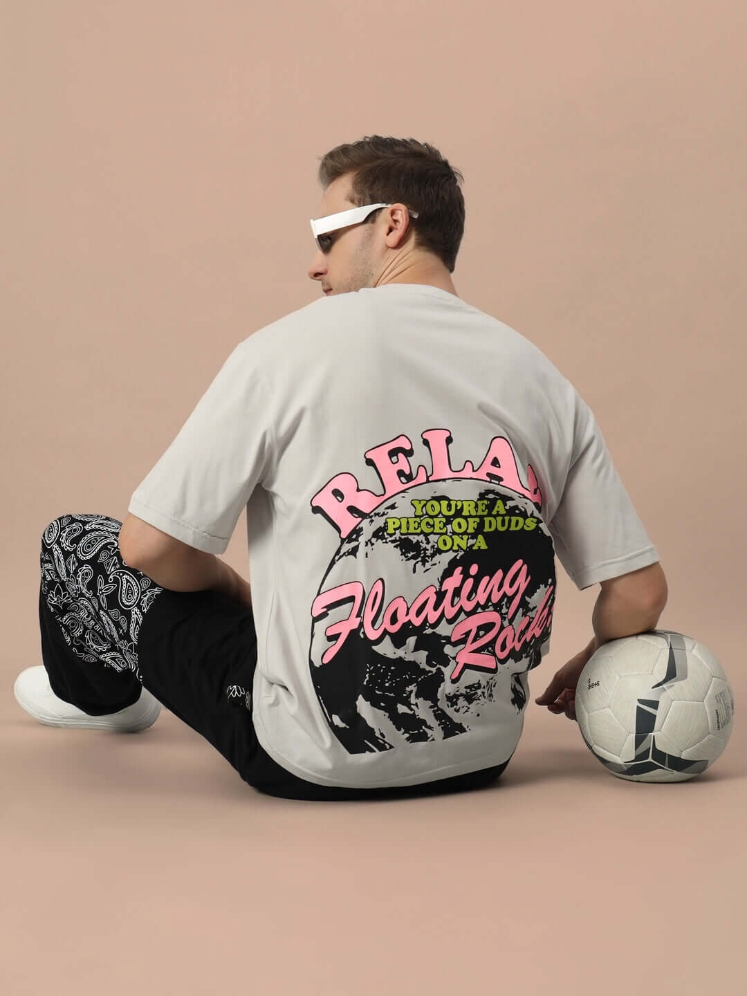 FLOATING ROCK OVER-SIZED T-SHIRT GREY worn by model sitting with a soccer ball, featuring bold graphic text design on back.