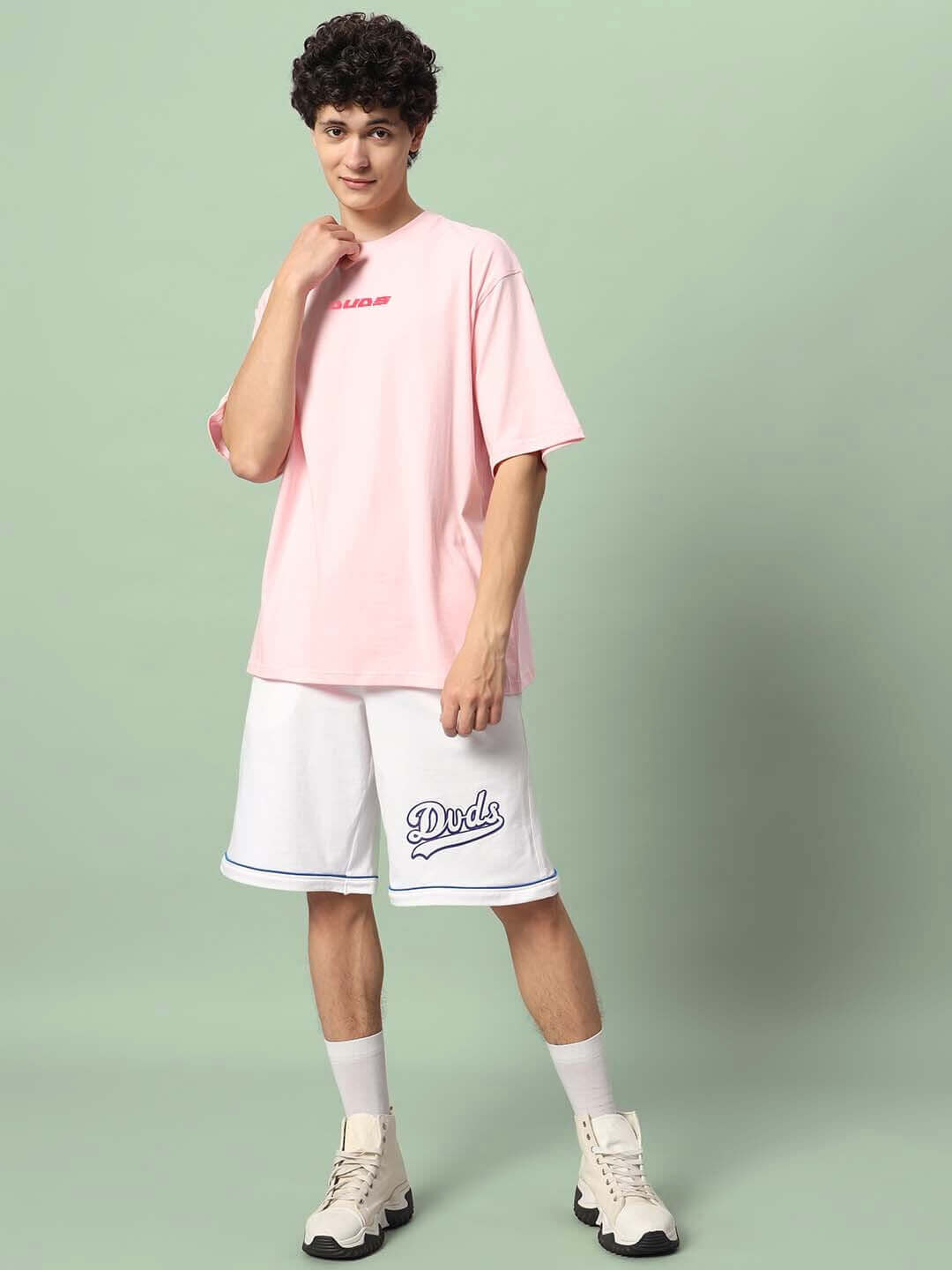Man wearing ILLUSION OVER-SIZED T-SHIRT PINK with white shorts standing against a green background.