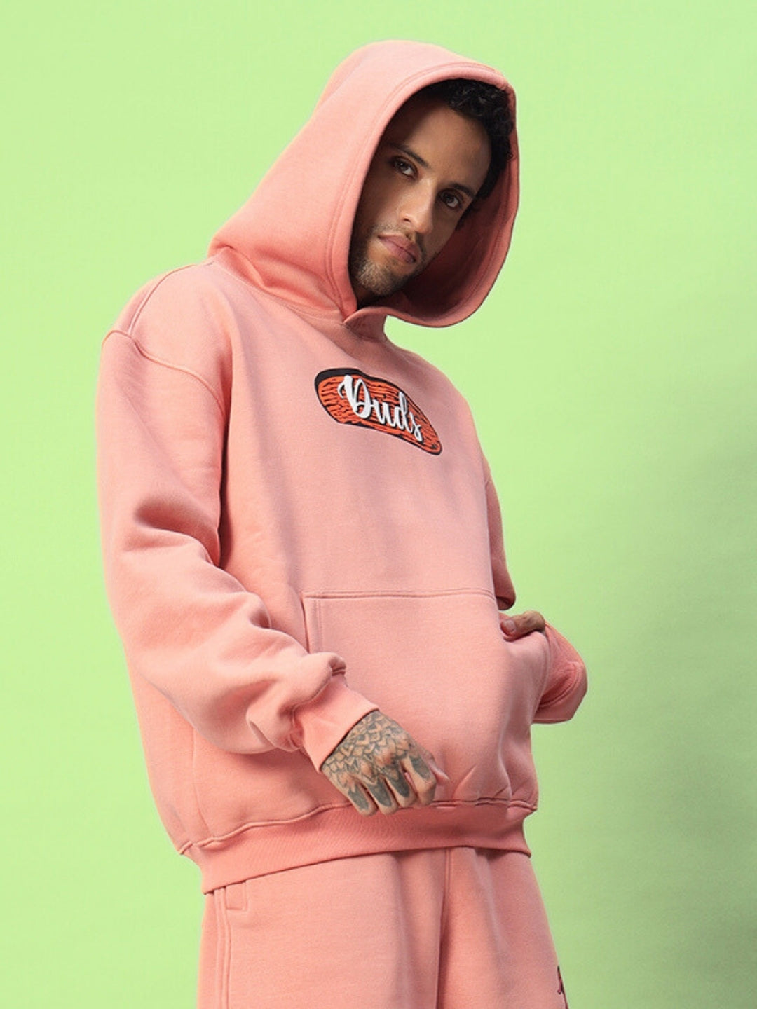 Model wearing Deliberate Fleece Hoodie in peach color with front logo, showcasing relaxed fit and cozy design against green background.