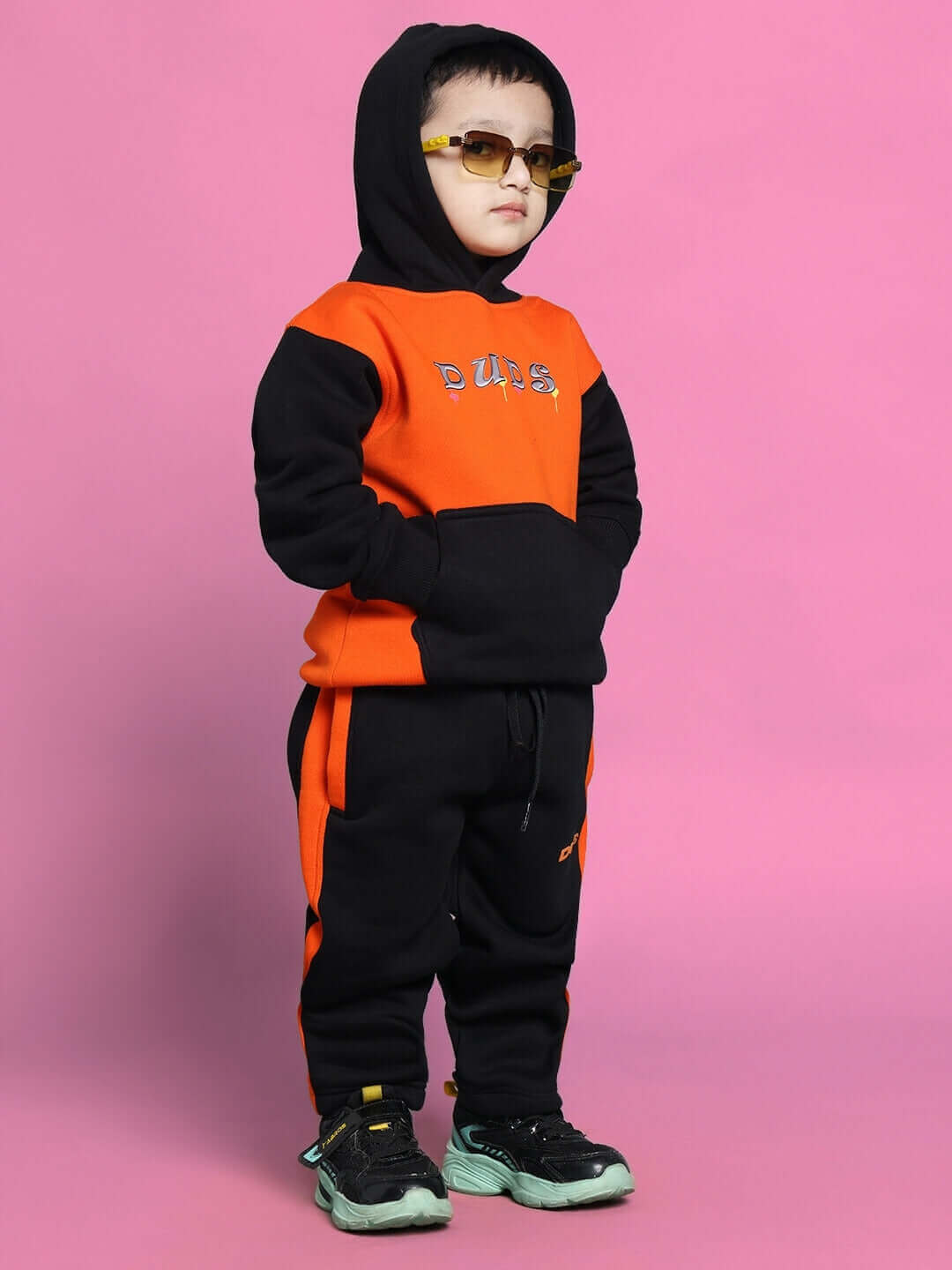 Young child wearing a black and orange COOL TEDDY co-ord, standing confidently against a pink background with sunglasses.