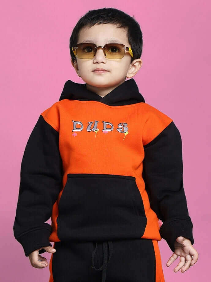 Child wearing COOL TEDDY CO-ORD for boys and girls in black and orange, stylish hooded sweatshirt and pants, against pink background.