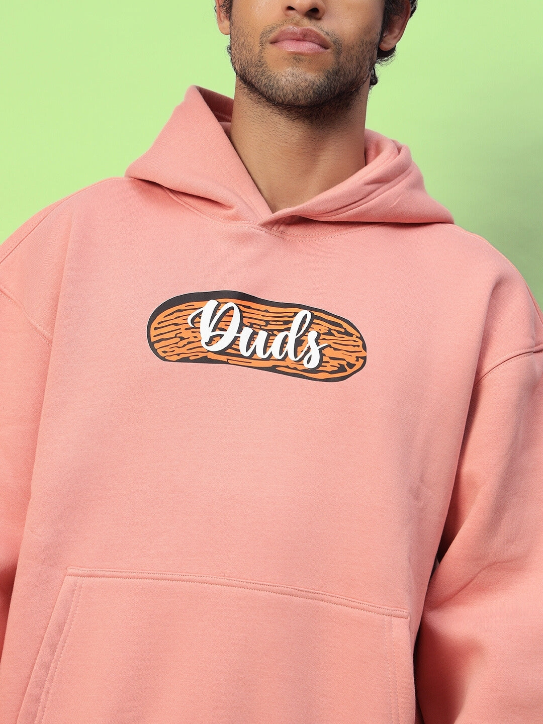 Man wearing Deliberate Fleece Hoodie in peach with graphic text, featuring a relaxed fit and front pocket, set against a green background.