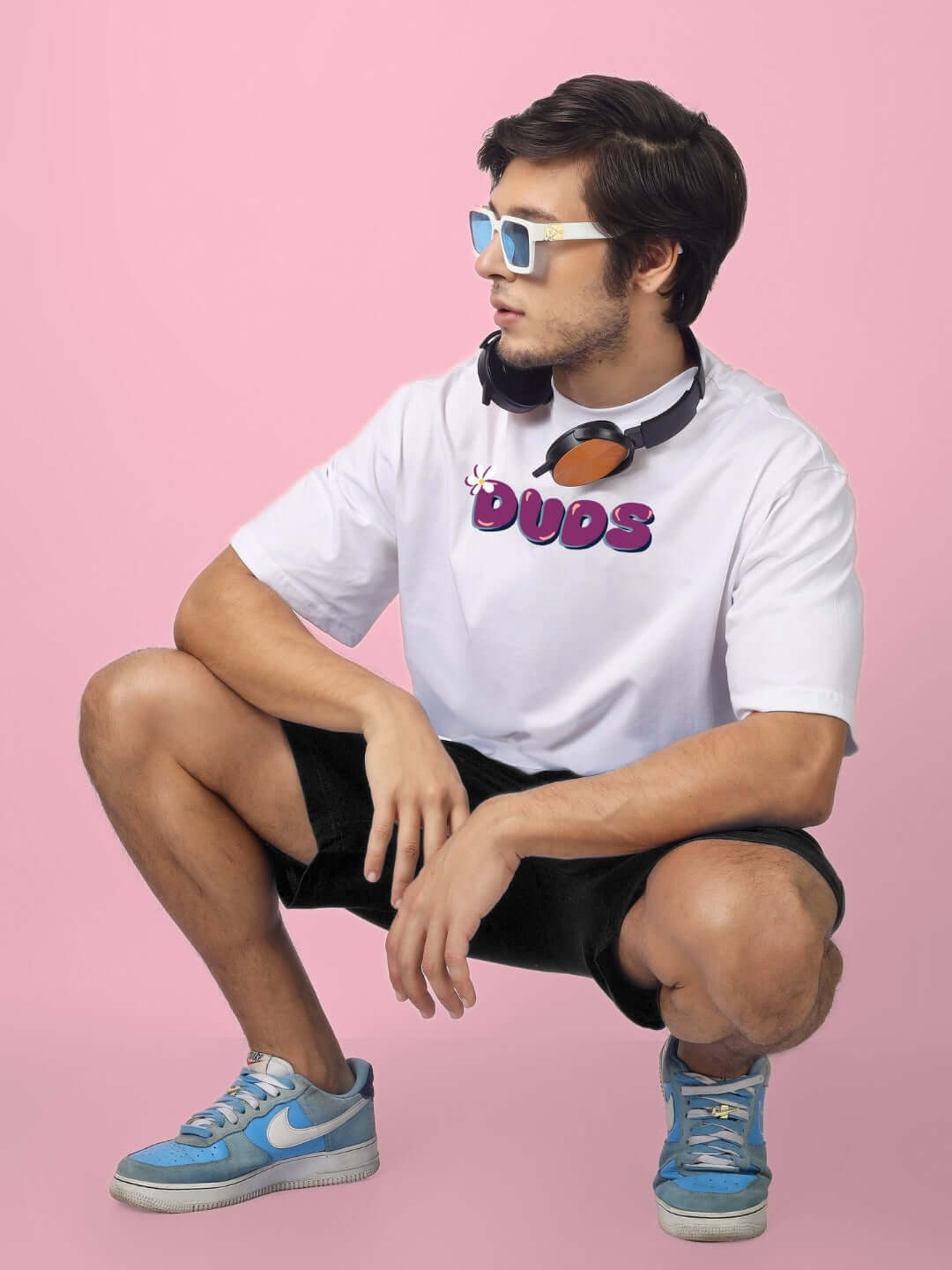 Man wearing a white graphic t-shirt, black shorts, sunglasses, and blue sneakers, posing on a pink background with headphones around his neck.